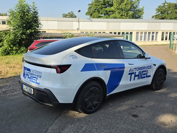 Weißer Tesla mit blauer Beschriftung „Autoservice Thiel“ auf dem Werkstattgelände
